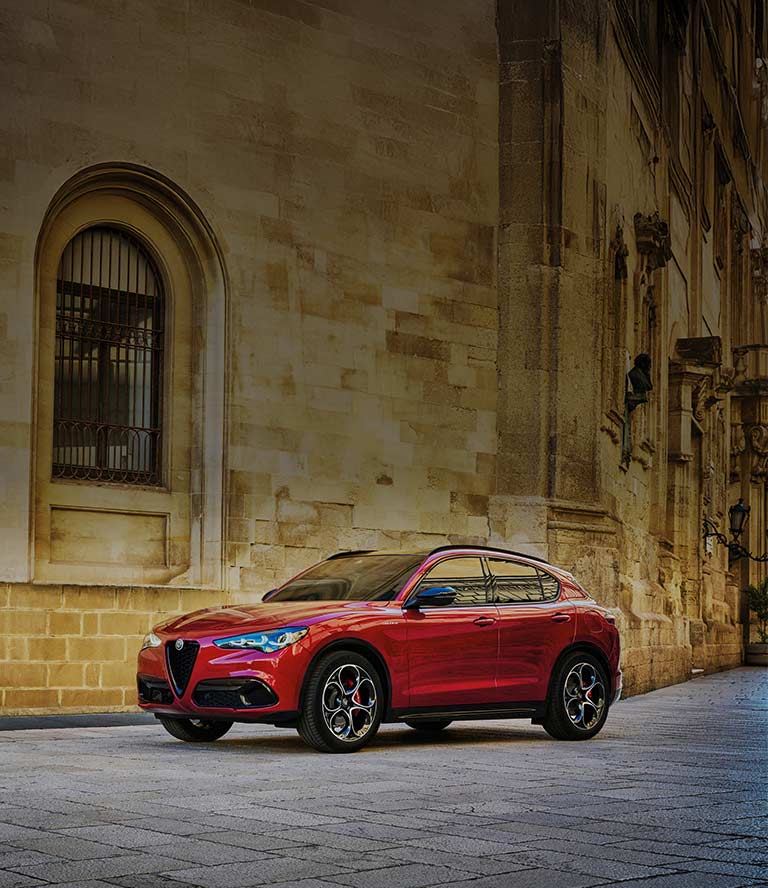 A driver-side front angle of a red 2026 Alfa Romeo Stelvio Veloce parked beside a brick wall.