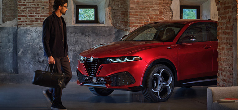 Un Alfa Romeo Tonale Sport Speciale 2026 rojo con los faros delanteros encendidos, estacionado entre dos edificios de ladrillo, con un hombre de pie frente al vehículo.