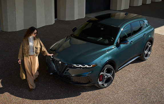 A raised angle of a green 2026 Alfa Romeo Tonale Sport Speciale parked beside a commercial building with a woman standing, touching the hood.