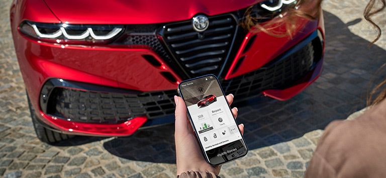 Vista frontal de un Alfa Romeo Tonale Sport Speciale 2026 rojo junto a una mujer que sostiene un smartphone con la aplicación My Alfa Romeo en pantalla.