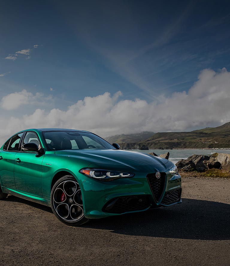 A passenger-side front angle of a green 2026 Alfa Romeo Giulia Veloce parked on a rocky beach, with ominous storm clouds in the background.