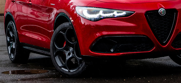 The lower portion of the front and passenger side of a red 2026 Alfa Romeo Giulia Veloce, including the wheels tires, as it travels in the rain.
