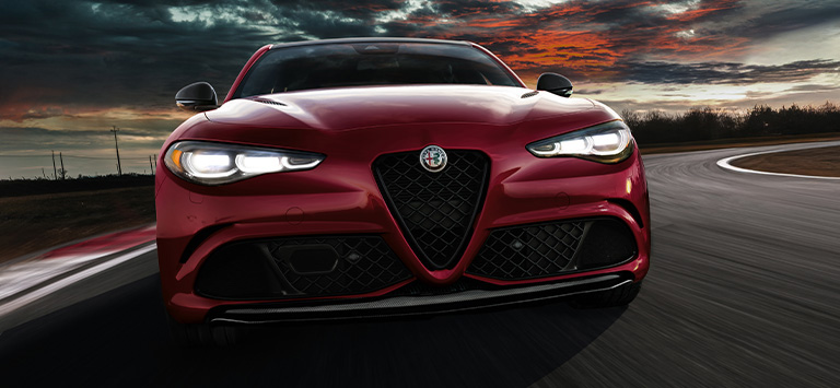 A front angle of a red 2026 Alfa Romeo Giulia Veloce with its headlamps on, traveling down a curved track at dusk.