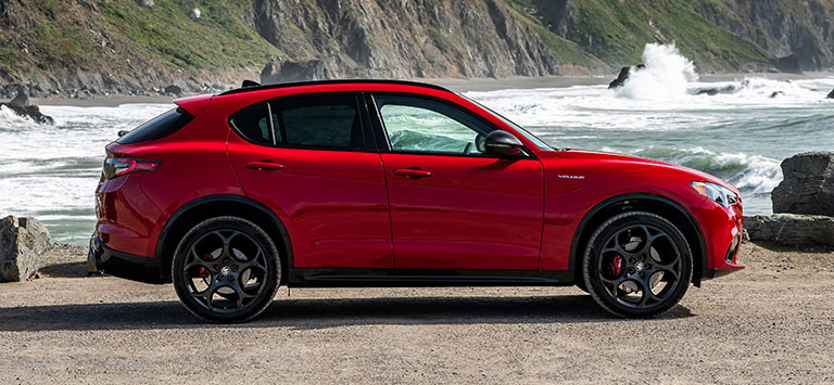 A passenger-side profile of a red 2026 Alfa Romeo Stelvio Veloce parked on a rocky beach.
