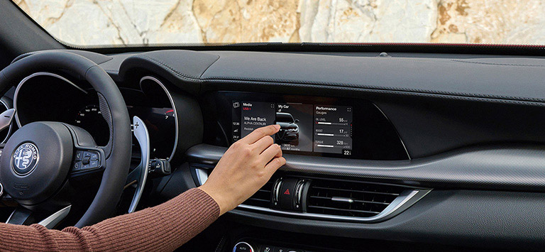 The steering wheel, Uconnect touchscreen and heating and cooling vents in a 2026 Alfa Romeo Stelvio with the driver touching the screen.