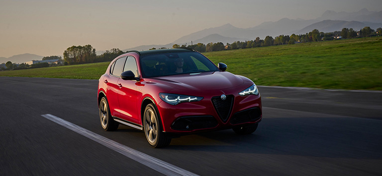 A red 2026 Alfa Romeo Stelvio Veloce with its headlamps on, traveling down a highway at dusk.